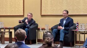 U.S. Sen. Michael Bennet, left, and Attorney General Phil Weiser sit on chairs holding mircophones.