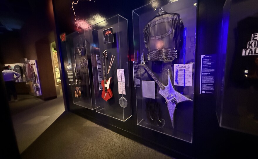 Three glass museum cases on a black wall show off music memorabilia, including a black, red and silver guitar, a leather vest and pants, a metal-studded leather jacket, drumsticks, a drumhead and four handwritten pieces of paper.