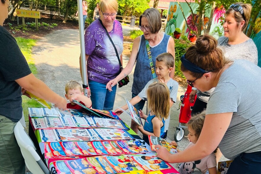 Several families with small children picking up books from a table outside at an event.