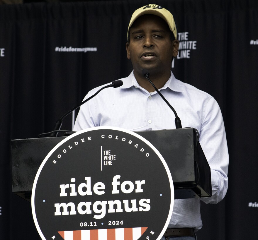 A middle-aged black man speaks at a podium with the words "ride for Magnus" printed on it