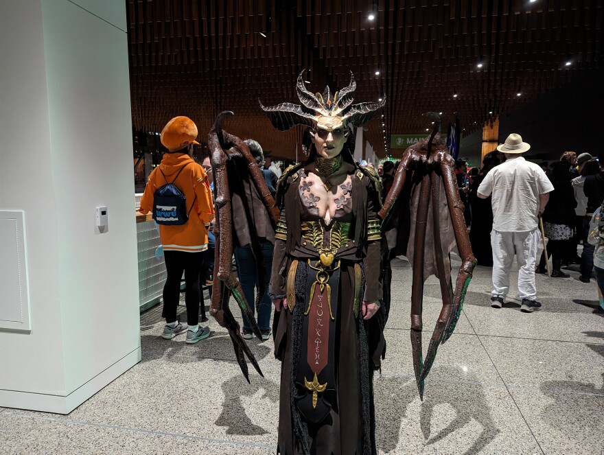 A person wears a costume with leather-like wings with a multi-layered robe, face paint and a four horned headpiece.