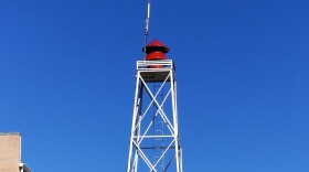 A white, vertical tower with a red siren on top. A blue sky fills the background of the composition.