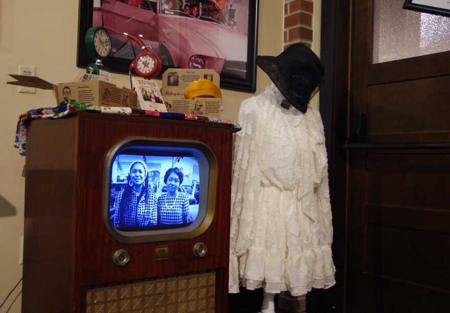 Two girls or young women can be seen on a old black and white TV. Assorted pictures, models of old gas pumps and more can be seen at the top of the TV. Next to it is a headless mannequin displaying a elegant white dress, with a black hat hanging on top.