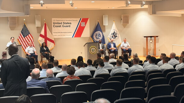 Coast guard graduates sit in the front two rows as an office speaks from a podium on the stage. 