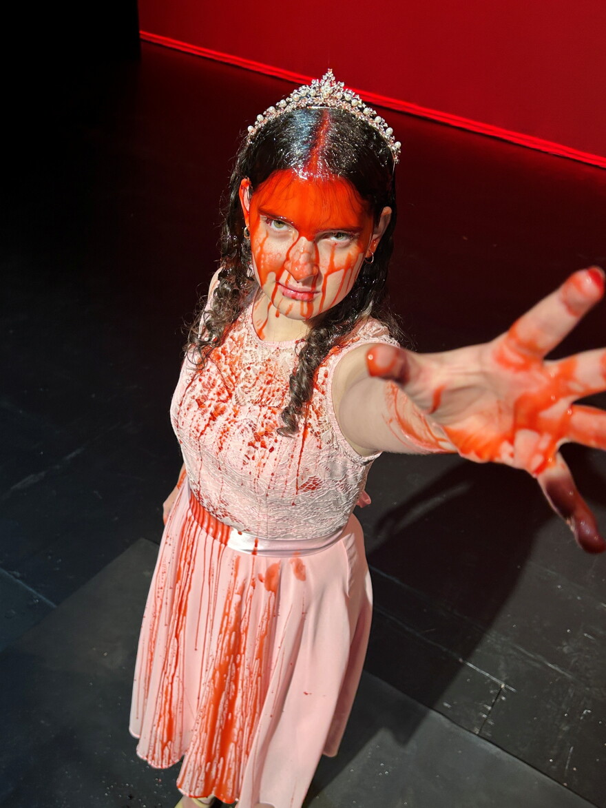 Young woman in pink dress wearing crown covered in fake blood looks up to the camera reaching her bloody hand up 