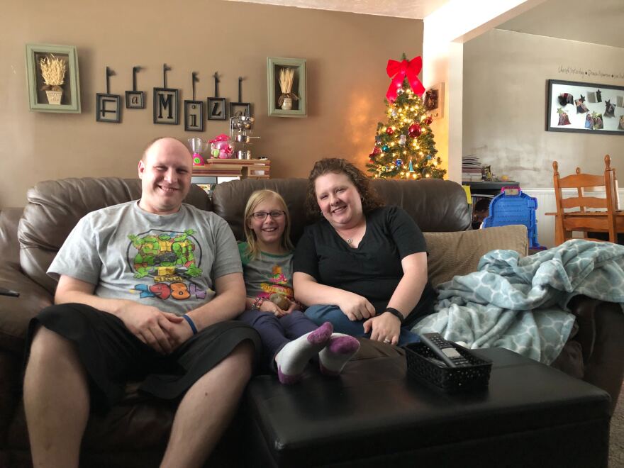 A man, child and woman sit smiling on a sofa. Behind them is a Christmas tree and a sign that says "family."