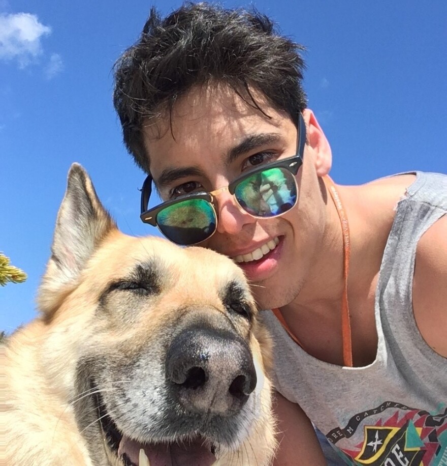 A man wearing mirrored sunglasses and a gray tank top looks down into the camera beside a light-furred and smiling dog.