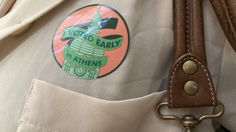 A close-up of an "I voted early in Athens" sticker on someone's beige blouse, next to the brown leather straps of their handbag. 
