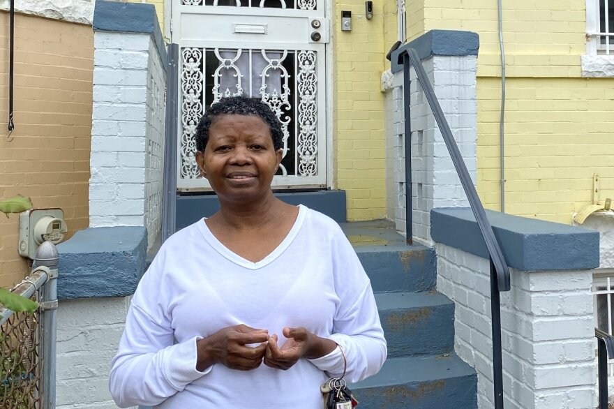 A woman wearing a white long-sleeved shirt stands half-smiling in front of a yellow house.