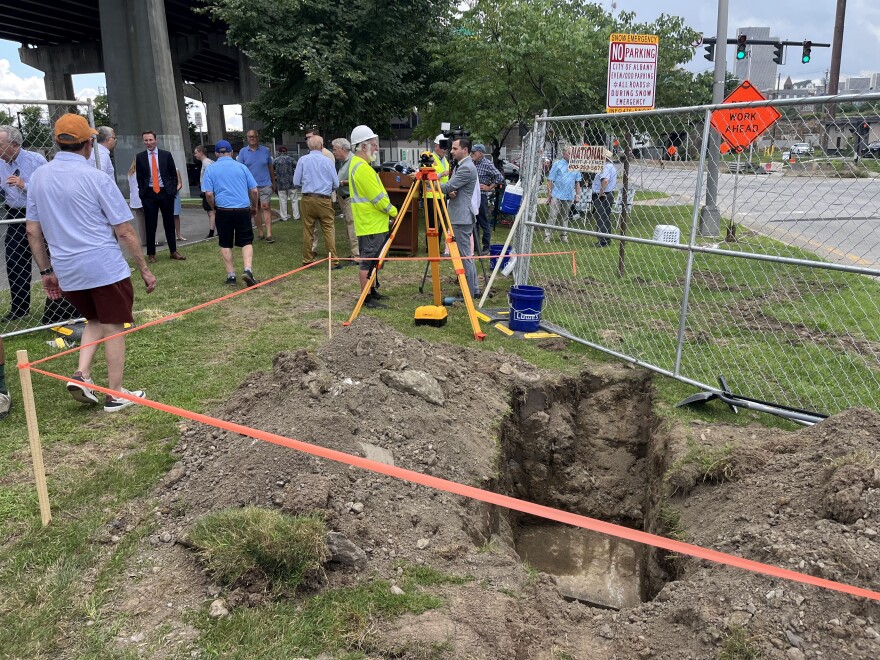 This is a photo of a grass patch between a parking lot and a road (peak Upstate New York). There's an approximately two-foot hole in the ground with piles of dirt on other side. The hole is blocked in by orange tape on one side and chain link fence on the other. There's a yellow tripod and a blue bucket next to the hole. A crowd of people is standing in the background next to a dig site. One of them is dressed in a hard hat and neon vest and is being interviewed by a television news reporter. 