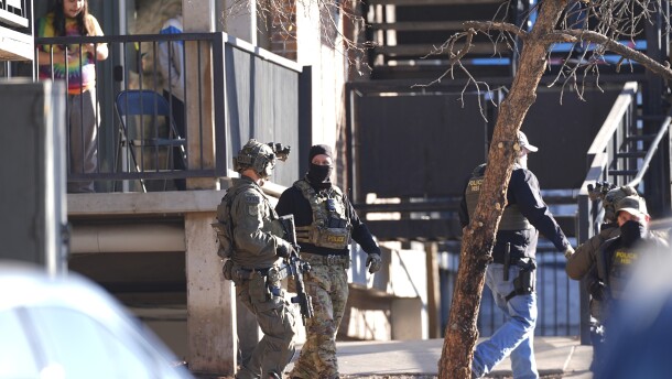 Multiple people in camouflage and tactical gear carry rifles outside of a large building.
