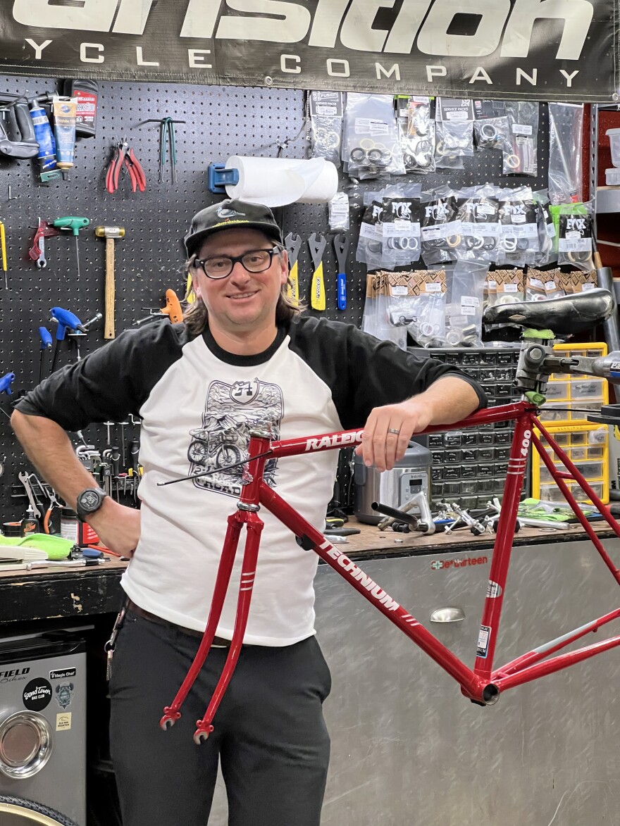 A man with glasses and a hat leans on a bike frame being held in a bike stand. A plethora of tools and supplies hangs behind him on a wall.