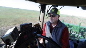 A white man inside a green tractor with grey facial hair, wearing black sunglasses, a green and yellow trucker hat, a red long sleeve shirt with a grey vest over. Farm land can be seen through the windows behind the man. 