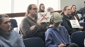 Two people seated in a crowded meeting space holding signs that read 'Data Centers' with a X over it and 'No Data Centers'
