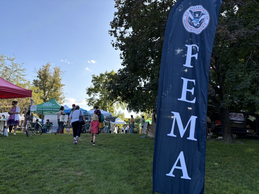 A blue flag with the letters FEMA stands in front of a farmers market where people are walking around