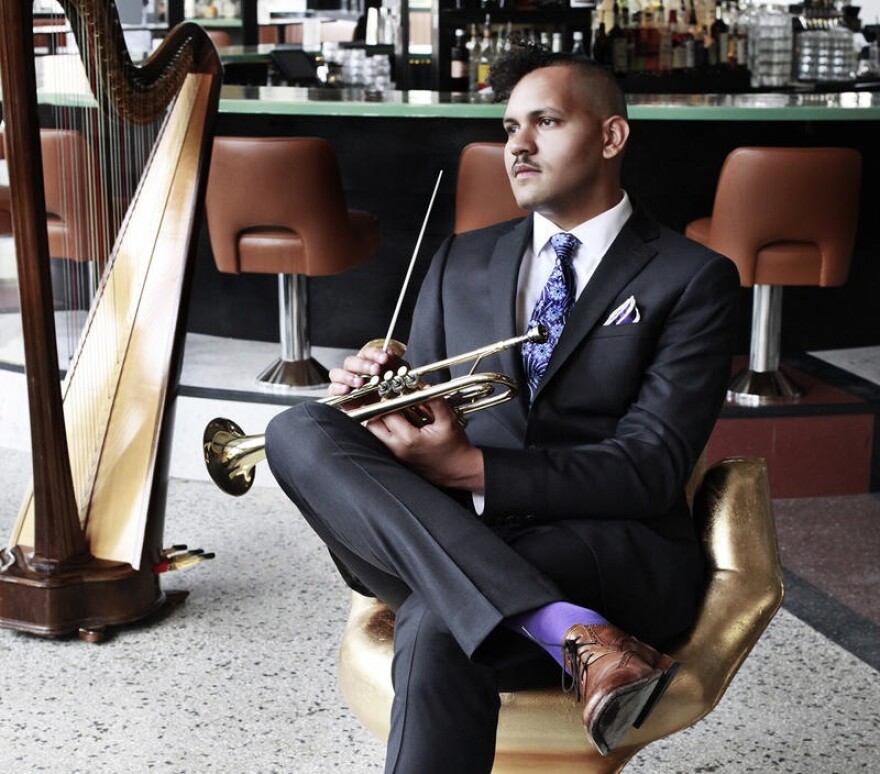 A Black man in a dark suit sits holding a trumpet. There is a harp to his right.