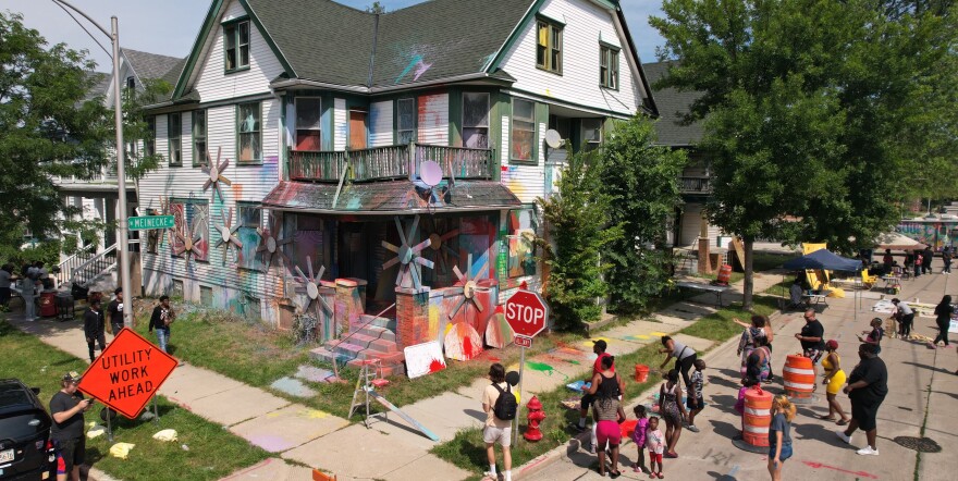 People surround a two story house that is covered with splashes of color. 