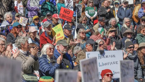 A crowd of protestors clap hands and hold signs that voice displeasure with President Donald Trump's governing practices.