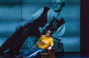 A man on stage in a yellow t-shirt and wearing a gold crown is stretched on a rock, with a projection of a man in Shakespearean style clothing looming behind him.