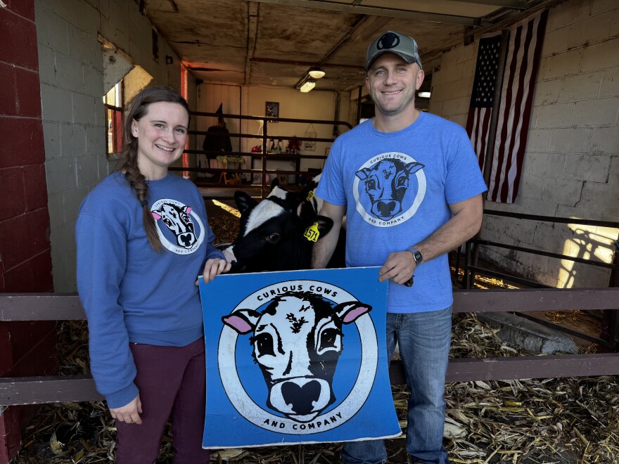 A woman on the left side of the image holds a sign with a cow in the middle and the words "Curious Cows And Company" written on the edges. To the right of the image, a man holds the same sign. A cow approaches the pair from behind at their barn.