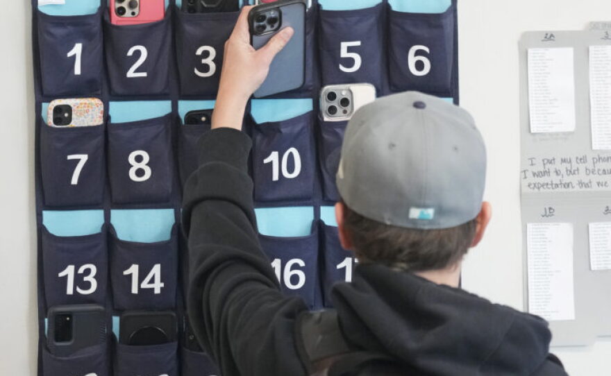 A ninth grader places his cellphone into a phone holder as he enters class at Delta High School, Friday, Feb. 23, 2024, in Delta, Utah. At the rural Utah school, there is a strict policy requiring students to check their phones at the door when entering every class. Each classroom has a cellphone storage unit that looks like an over-the-door shoe bag with three dozen smartphone-sized slots. 