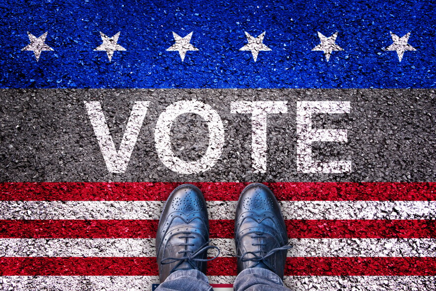 Feet on and asphalt road with the word vote. Red and white stripes are below the word. White stars on a blue stripe are above.