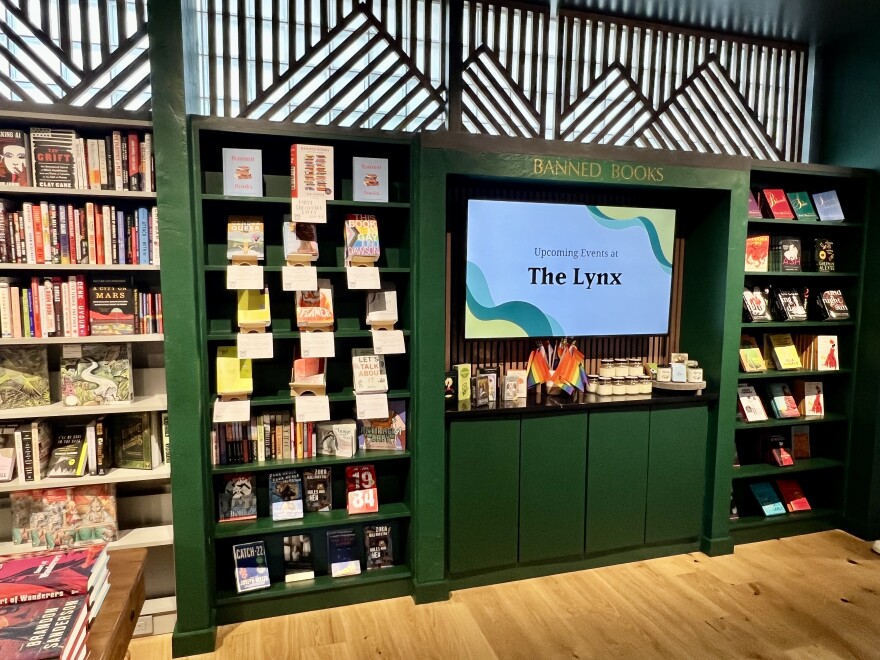 A dark green bookshelf filled with books. On the top of the bookshelf reads "Banned Books" in gold letters. In the middle of the shelf is a TV with a screen that reads "Upcoming Events at The Lynx." In front of the TV are pride flags and candles.