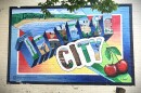 The image shows a post-card style mural paint on a beige brick wall that says “Greetings from Traverse City” in giant 3D block letters. The lettersT are filled with detailed illustrations of local features, including a lighthouse, sailboats, sand dunes, wine, and ski slopes. In the top right corner a pair of giant glossy red cherries sit. 