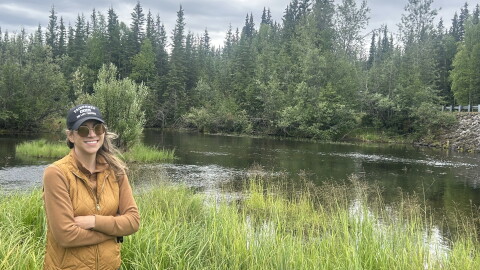 A woman in a fleece and cap stands beside a pond in a wilderness area.