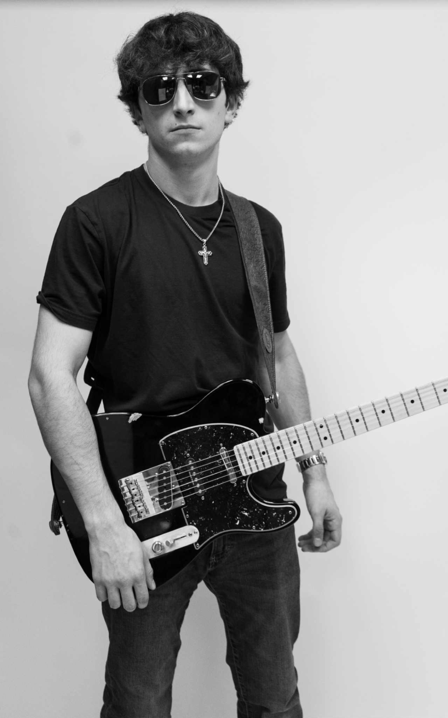 Black-and-white portrait of a young man wearing sunglasses, a T-shirt, and a cross necklace while holding a black electric guitar against a plain background.