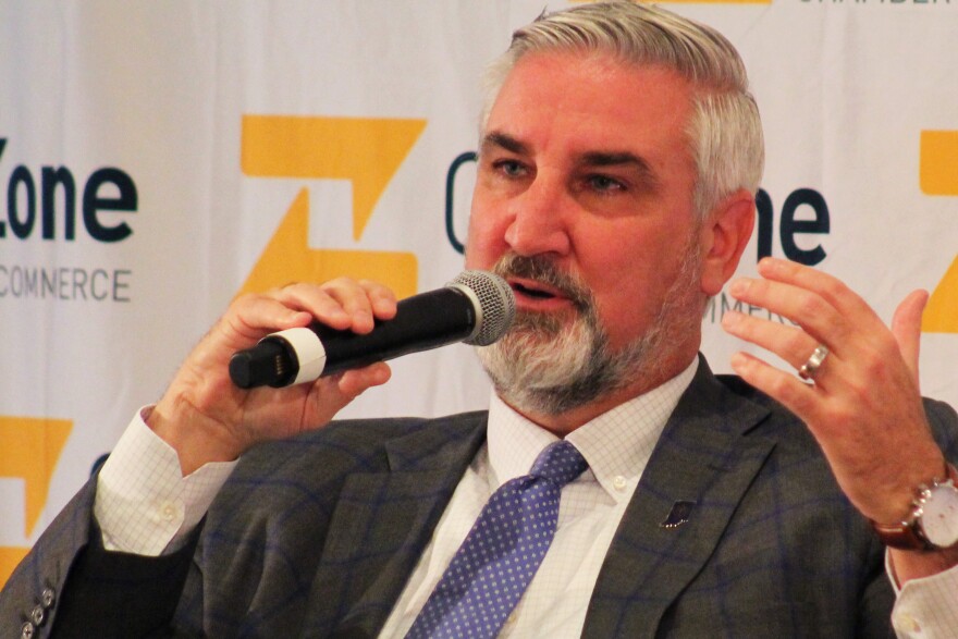 Indiana Governor Eric Holcomb sits, holding a microphone in his right hand and gesturing with his left, while speaking to a local chamber of commerce. Holcomb is a White man with white and gray hair and a beard. He's wearing a gray, checkered pattern suit with a white checkered shirt and blue and white polka dot tie.