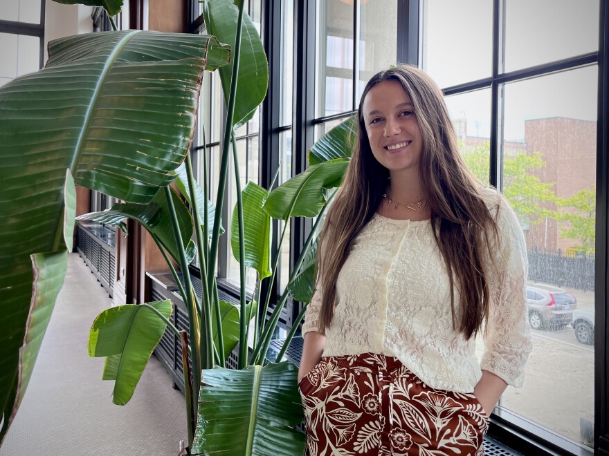 Kristen Plonsky wears an off-white shirt and patterned brown pants while she smiles for a photo beside a large green plant and in front of a black-paned window. Plonsky worked as a Health Hub Coordinator with Detroit Public Schools Community District. After the federal government increased immigration enforcement, her role shifted to helping local families navigate the immigration system and supporting them with basic needs.