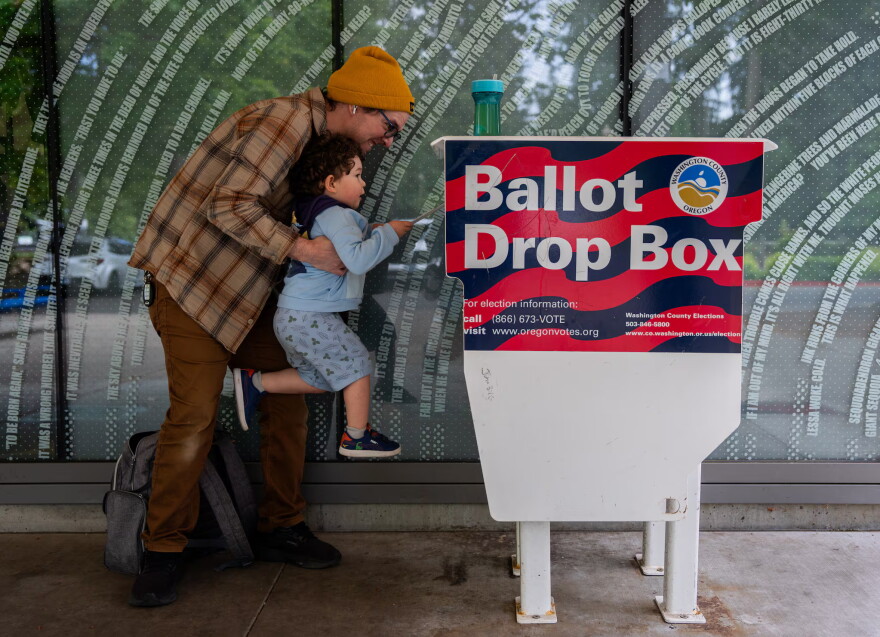 FILE - Matt lifts up his three-year-old son, Theo, to cast his ballot into the Hillsboro Public Library-Shute Park ballot drop box in May 2026. Election officials are increasingly recommending voters plan to use drop boxes rather than mailing their ballot.