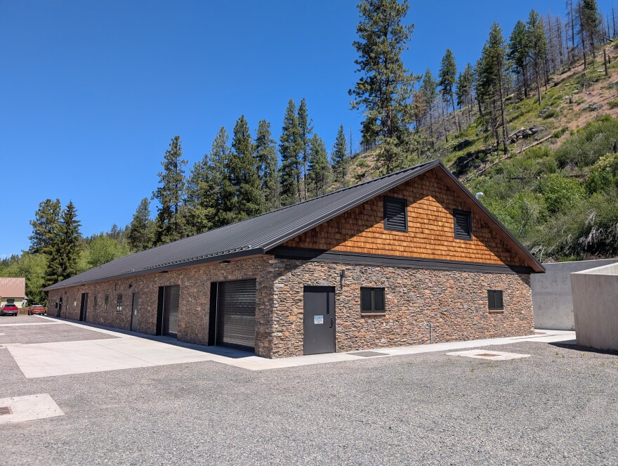 A long building with rock siding, a metal roof and wood shingles in the gables. The building is surrounded by concrete and gravel