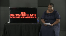 A woman sits to the right side of a sign that says "The Brown & Black Forums of America."