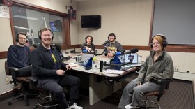 Five smiling people wearing headphones sit at a table in a radio talk studio: a man at left in foreground has short brown hair and a brown beard and is wearing glasses, a navy sweater and black pants; a man at right in foreground has short brown hair and a brown beard and is wearing a blue button-down shirt, blue and yellow t-shirt, black pants and white sneakers; a man at left in background has curly dark hair and is wearing a black t-shirt; a man at right in background has short dark hair and a dark beard and is wearing glasses and a grey sweatshirt; a woman at center has long red hair and is wearing a green sweatshirt, grey jeans and sneakers.