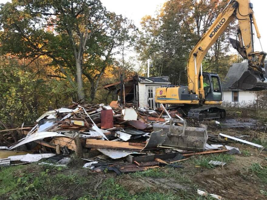 A yellow backhoe demolishes a small home. It's walls and windows have been torn apart.