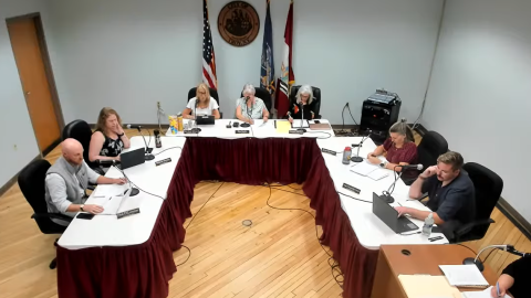 The seven members of the Troy City Council are sitting in a U-shaped arrangement of tables. There's some flags in the background. 