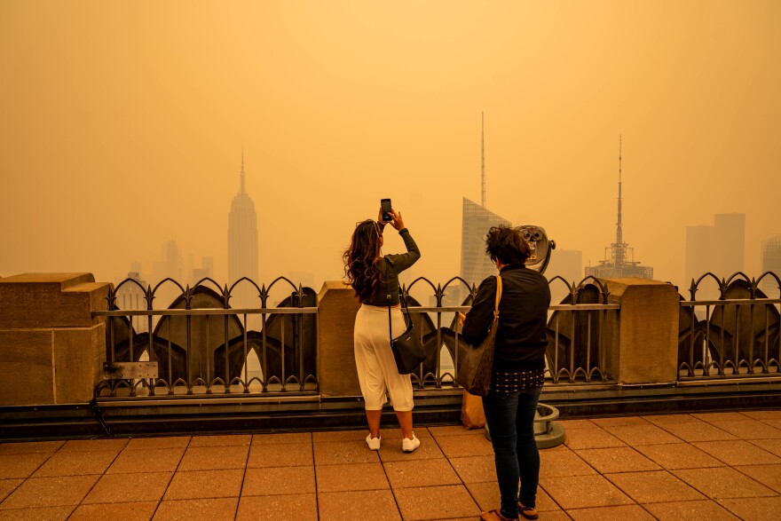 A woman takes a photo of a city skyline from a balcony with another woman standing behind her with smoggy orange-hued air.