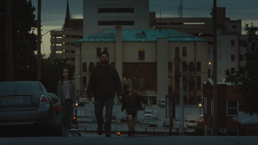 A middle aged man and two children walk down a street. The lighting is dark. 