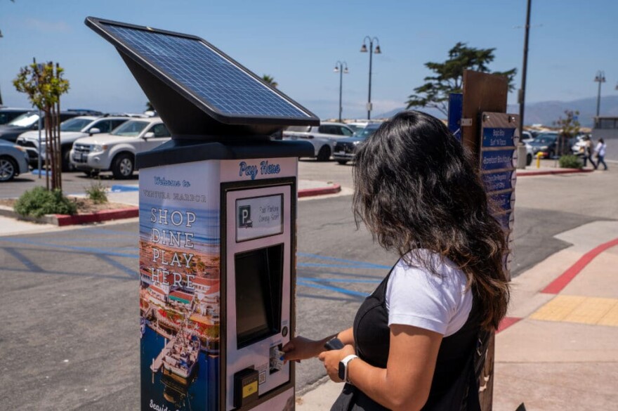 A woman stands at a pay kiosk in a parking lot. The kiosk has a built-in solar panel. Cars are parked in a lot in the background.