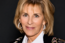 A smiling woman with short blonde hair, wearing white hoop earrings, a necklace, and a white blouse under a black blazer with hearts and stars embroidered on it.