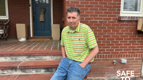 Michael Hickey sits outside his home. He wears a green striped shirt and jeans in front of a brick house.
