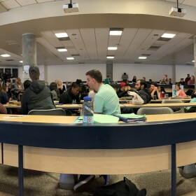 A panoramic photo. There are several rows of long tables with people sitting down and talking.