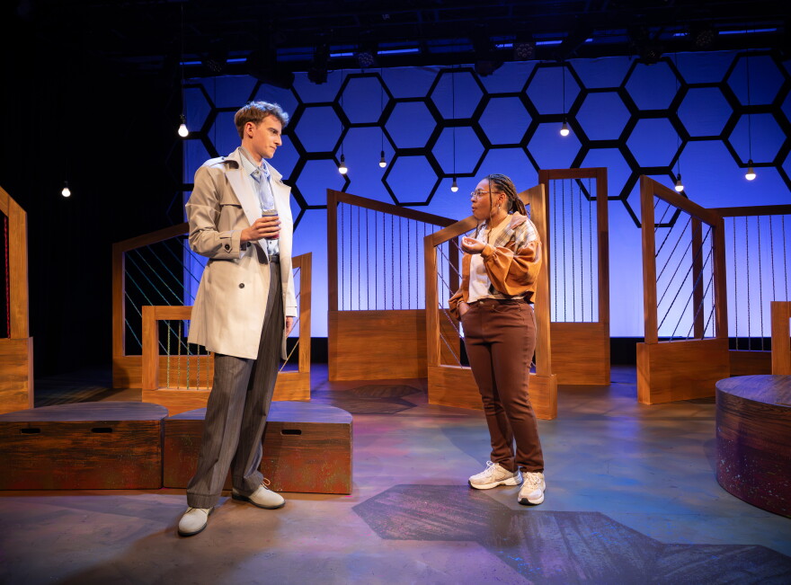 UGA students Jacob Arkin and Samara McKnight standing facing each other on a starkly dressed stage with brown, angled, wood and wire set pieces in front of a glowing blue-lit backdrop covered by the silhouette of a honeycomb pattern.