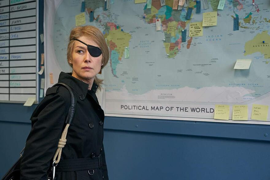 A blonde woman wearing an eyepatch and all black stands in front of a map with pins in it. 