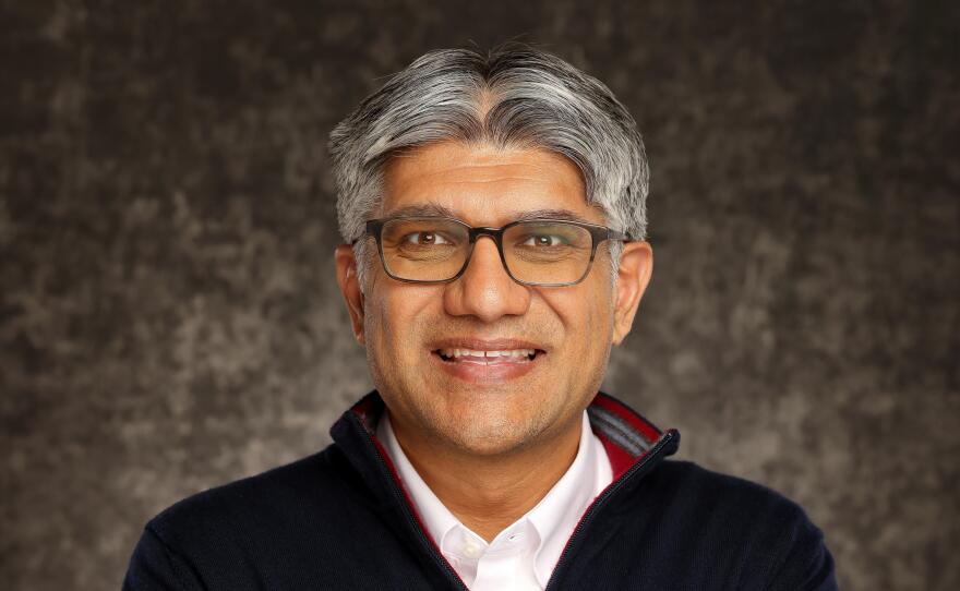A man with short grey hair is standing with his arms crossed and wearing a white button-down shirt under a navy sweater.