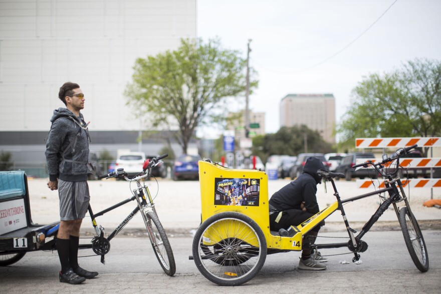 Two pedicab drivers waiting for rides. One driver is standing and the other is sitting on the passenger compartment.