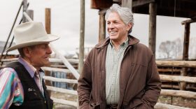 A man wearing a black vest and cowboy hat chats with another man wearing a coat. They're in front of corrals. 