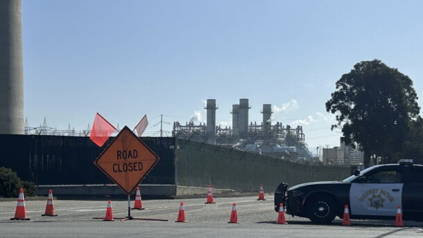 A road closed sign, police car, and traffic cones are shown in front of industrial smoke stacks. 